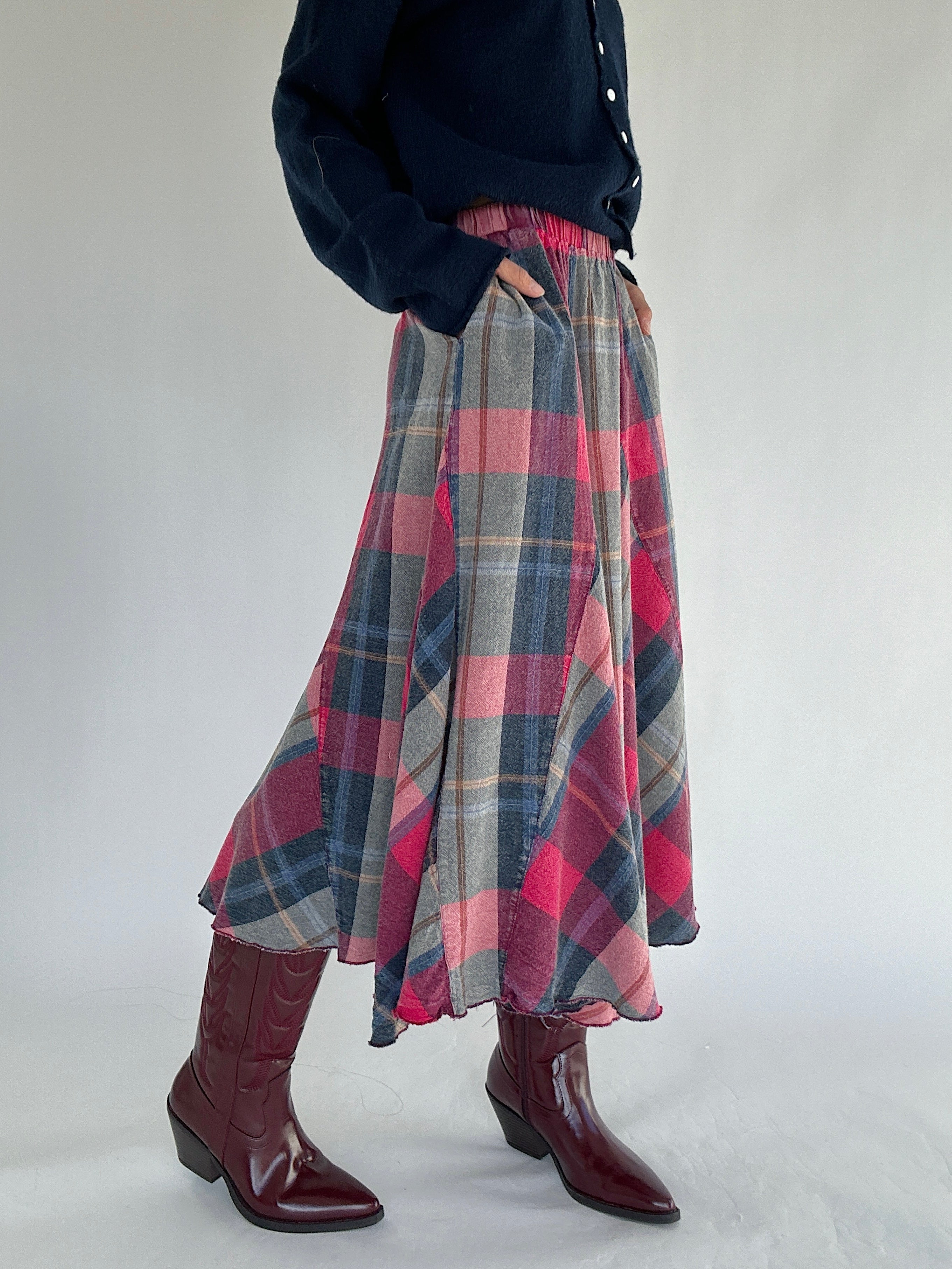 Person wearing a plaid skirt and nacy sweater with maroon boots on a light gray background showing skirt pockets