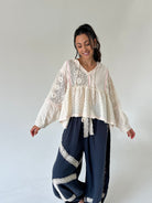 A woman modeling a bohemian-style outfit consisting of a cream lace and dot-print long-sleeve top paired with navy blue wide-leg pants featuring white lace trim, set against a plain light gray studio background.