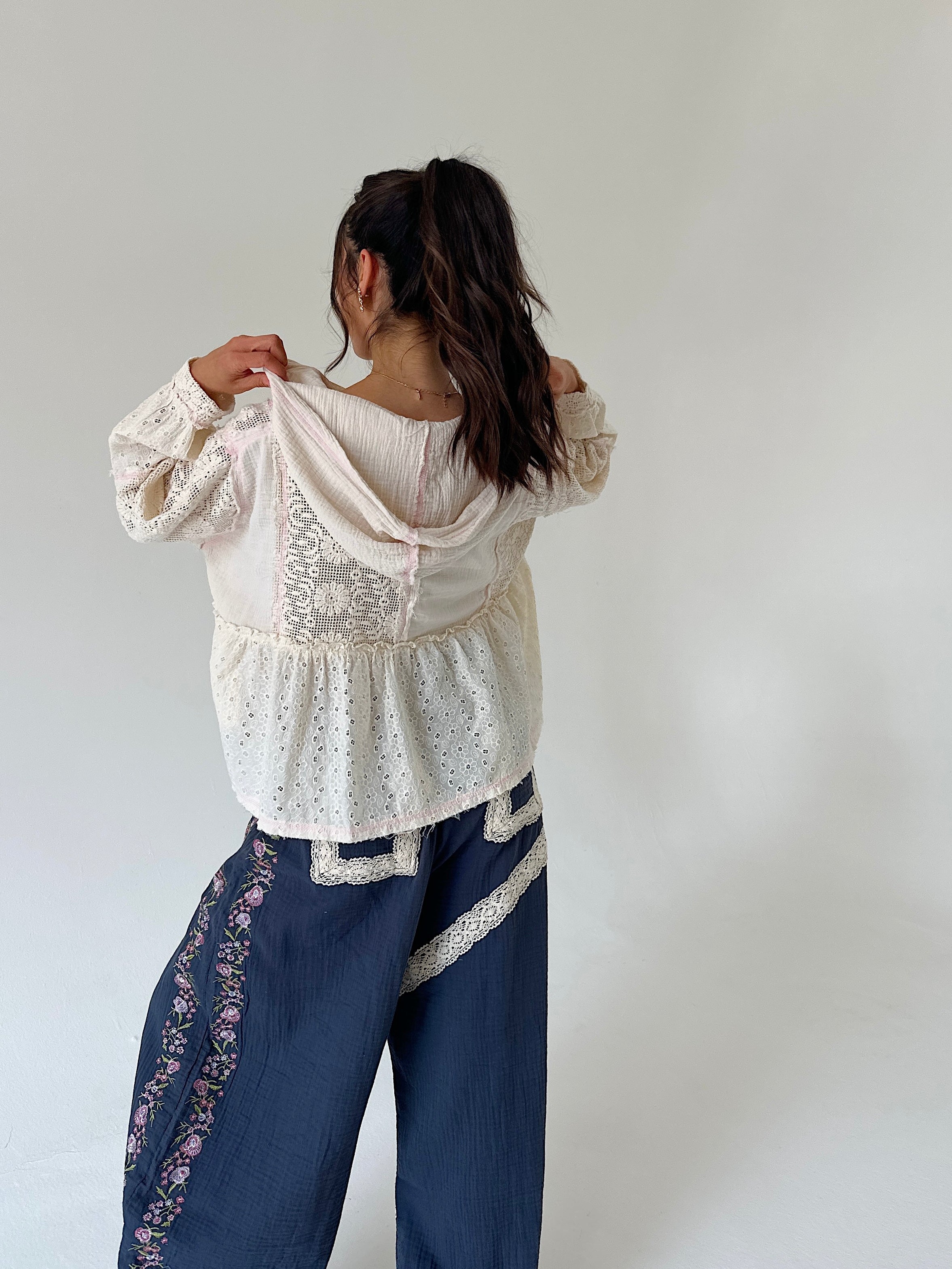 A woman shown from behind wearing a cream eyelet lace long-sleeve top and dark blue wide-leg pants with floral embroidery, posing against a plain light gray wall.