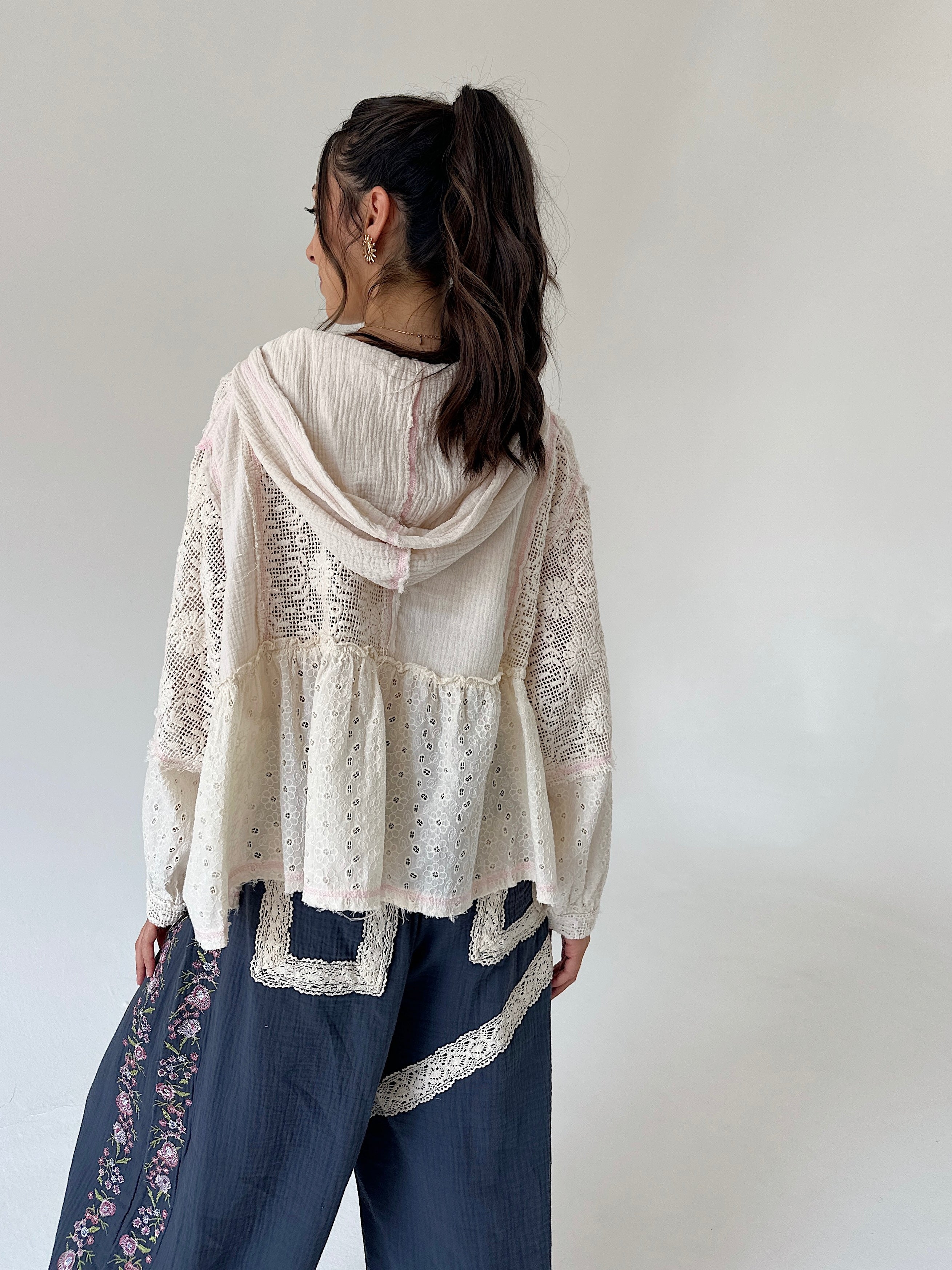 A woman shown from the back wearing a cream-colored lace and eyelet hooded top layered over dark blue pants with floral embroidery and lace trim, set against a plain light gray background.