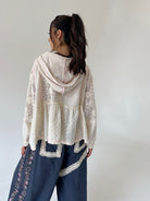 A woman shown from the back wearing a cream-colored lace and eyelet hooded top layered over dark blue pants with floral embroidery and lace trim, set against a plain light gray background.