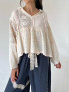 A woman wearing a cream eyelet lace long-sleeve blouse with a tie waist and dark blue wide-leg pants against a plain white background.