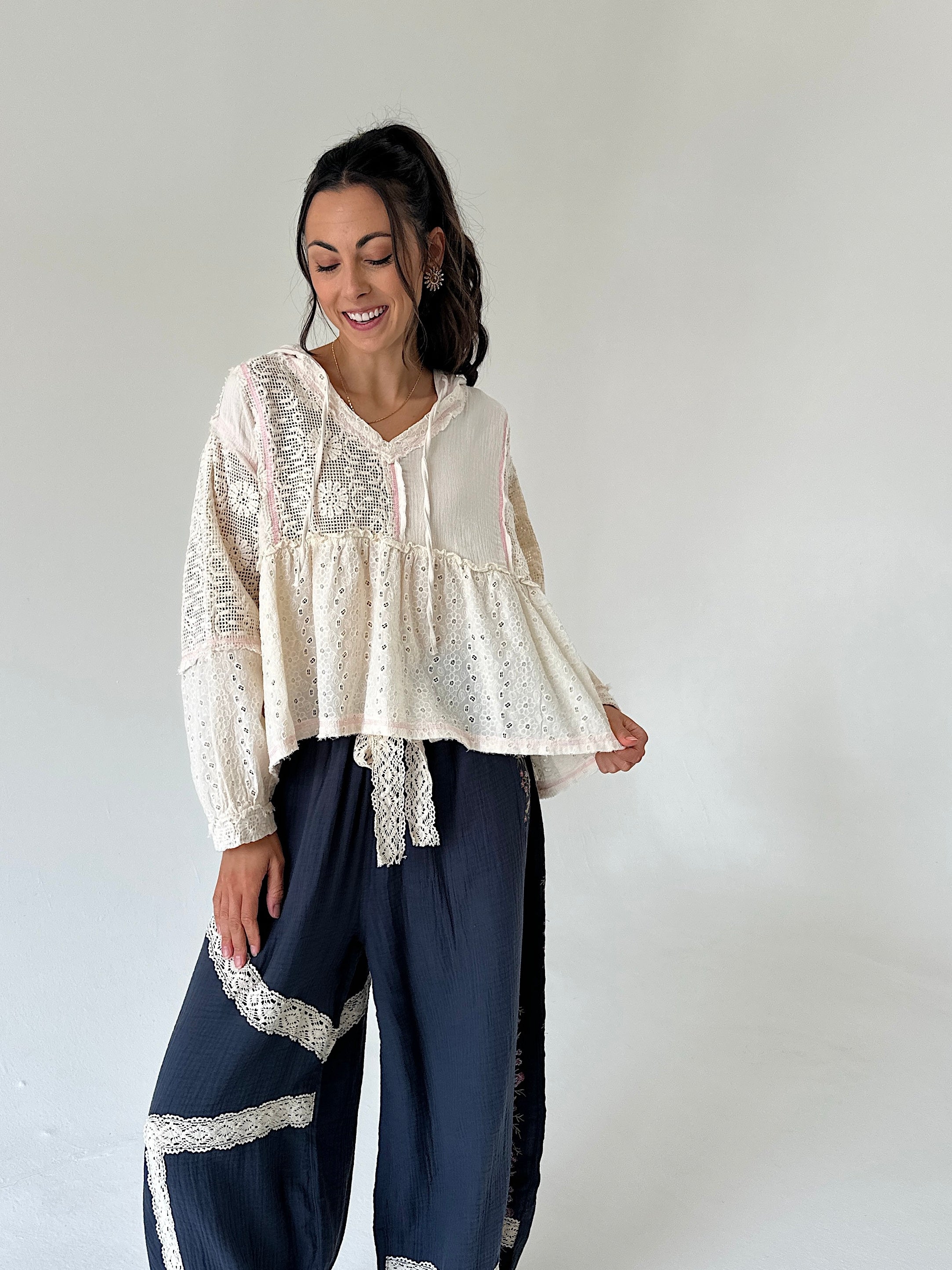 A woman modeling a cream-colored bohemian-style blouse with intricate eyelet embroidery and a ruffled peplum hem, paired with navy blue wide-leg pants featuring lace trim, set against a plain light gray studio background.