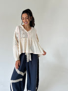A woman modeling a cream-colored bohemian-style blouse with intricate eyelet embroidery and a ruffled peplum hem, paired with navy blue wide-leg pants featuring lace trim, set against a plain light gray studio background.