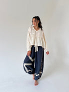 A woman stands barefoot in a studio wearing a cream lace long-sleeve top and navy blue wide-leg pants with white lace trim.