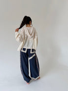 A woman stands barefoot against a plain light gray studio background, viewed from behind. She is wearing a cream lace long-sleeve top and loose dark navy wide-leg pants with cream trim and side floral embroidery.