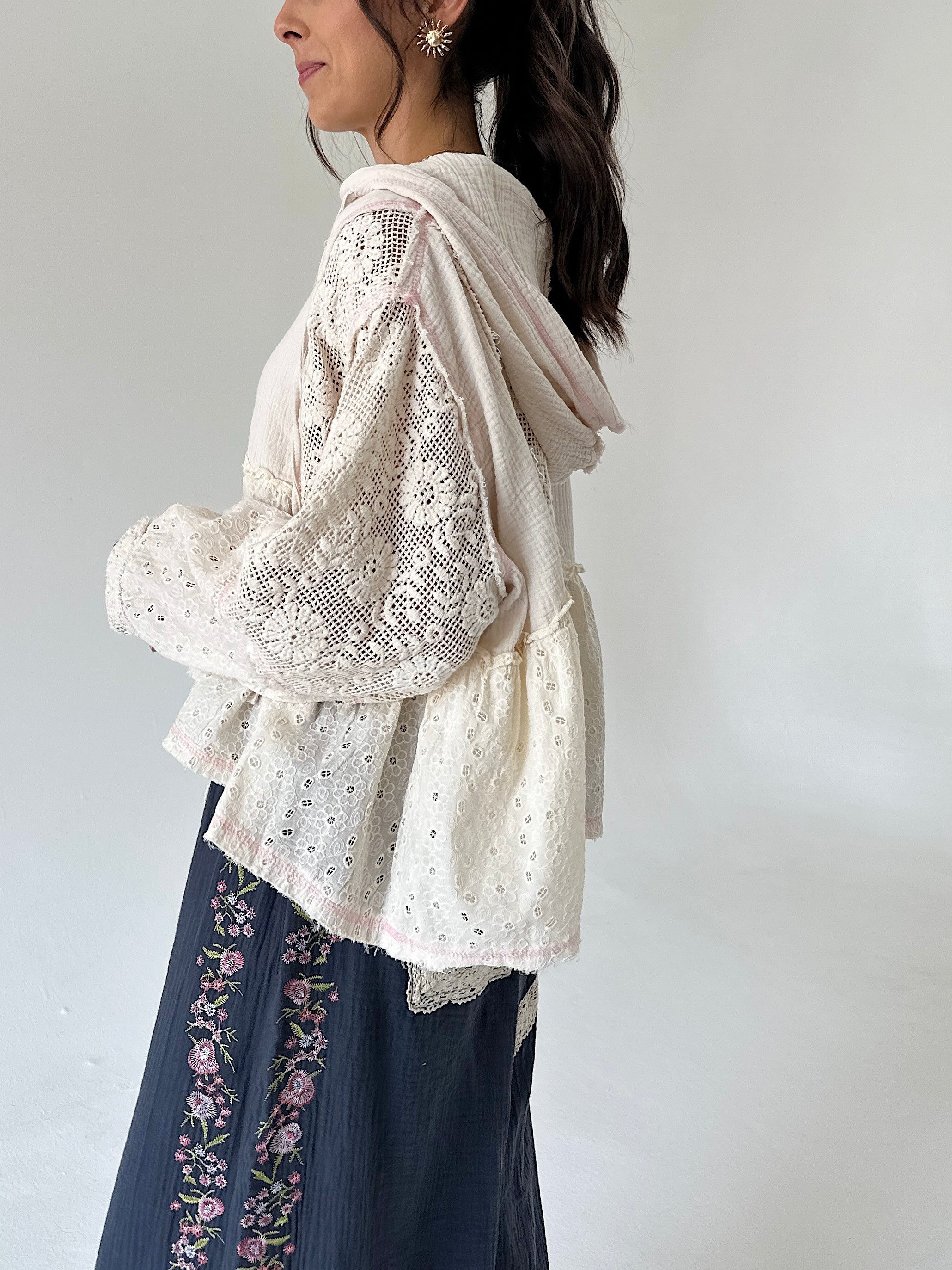 A woman shown from the side wearing a cream-colored eyelet lace long-sleeve hooded cardigan over a matching cream knit top, paired with dark blue floral embroidered pants, against a plain light gray background.