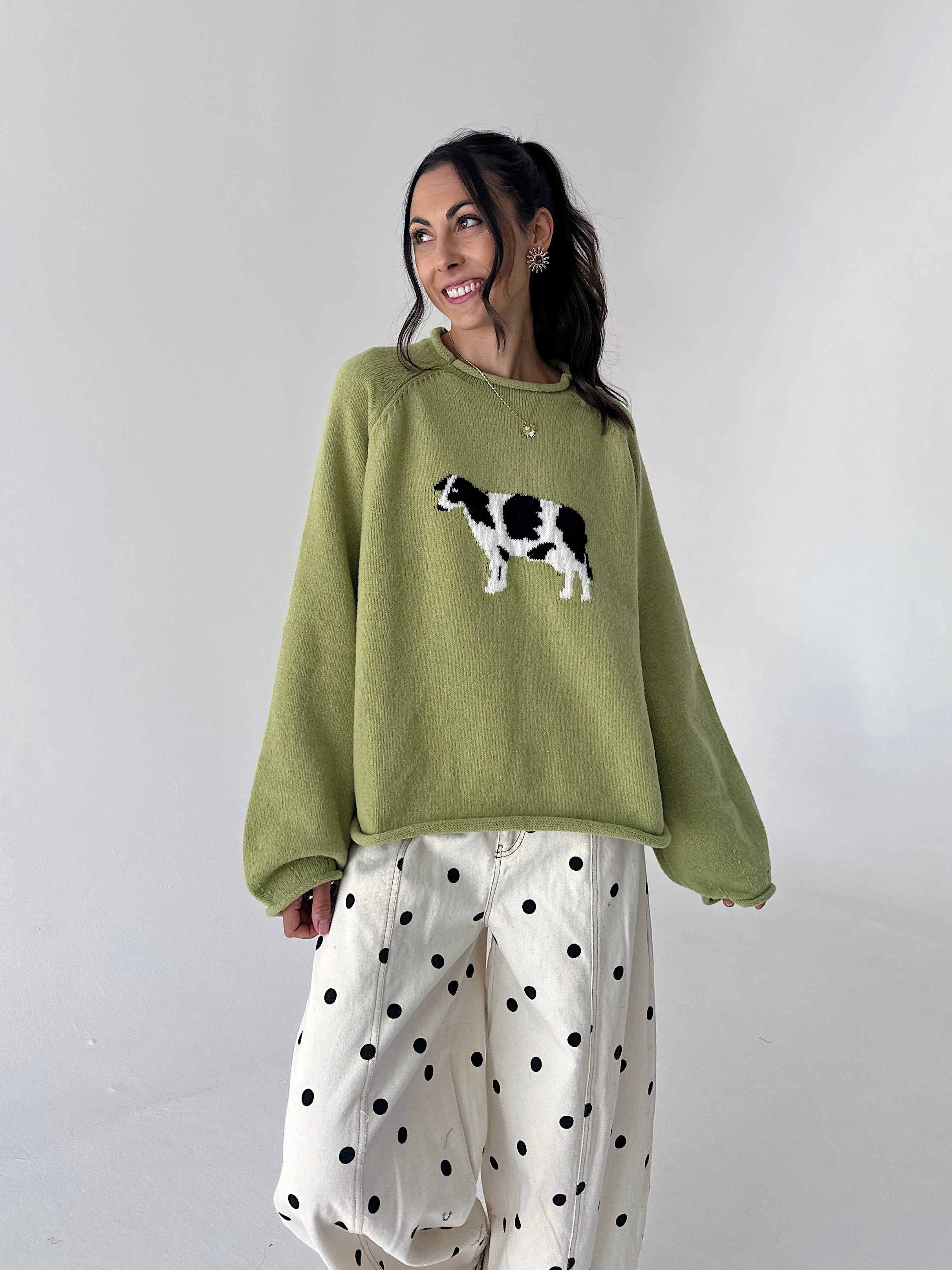 A woman wearing a sage green crewneck sweater with a black and white cow graphic on the chest, paired with white wide-leg pants featuring black polka dots, set against a plain light gray studio background.
