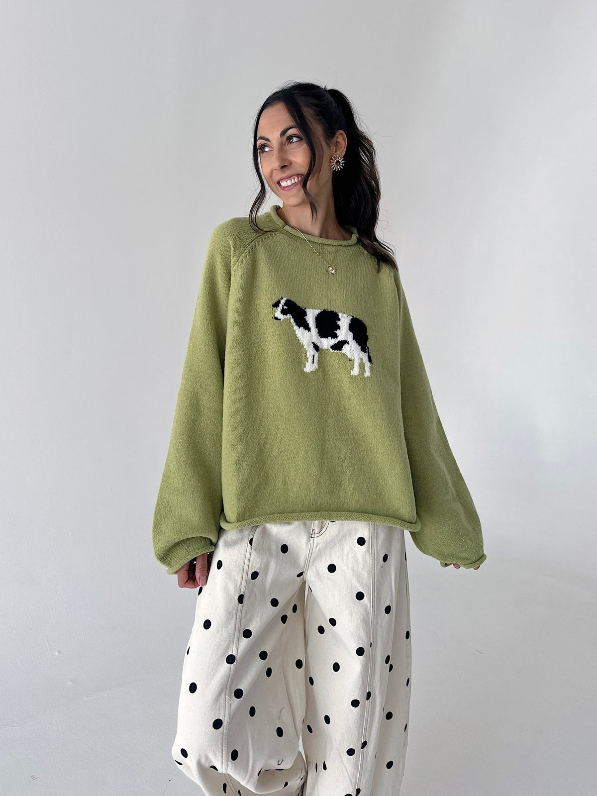 A woman wearing a sage green crewneck sweater with a black and white cow graphic on the chest, paired with white wide-leg pants featuring black polka dots, set against a plain light gray studio background.