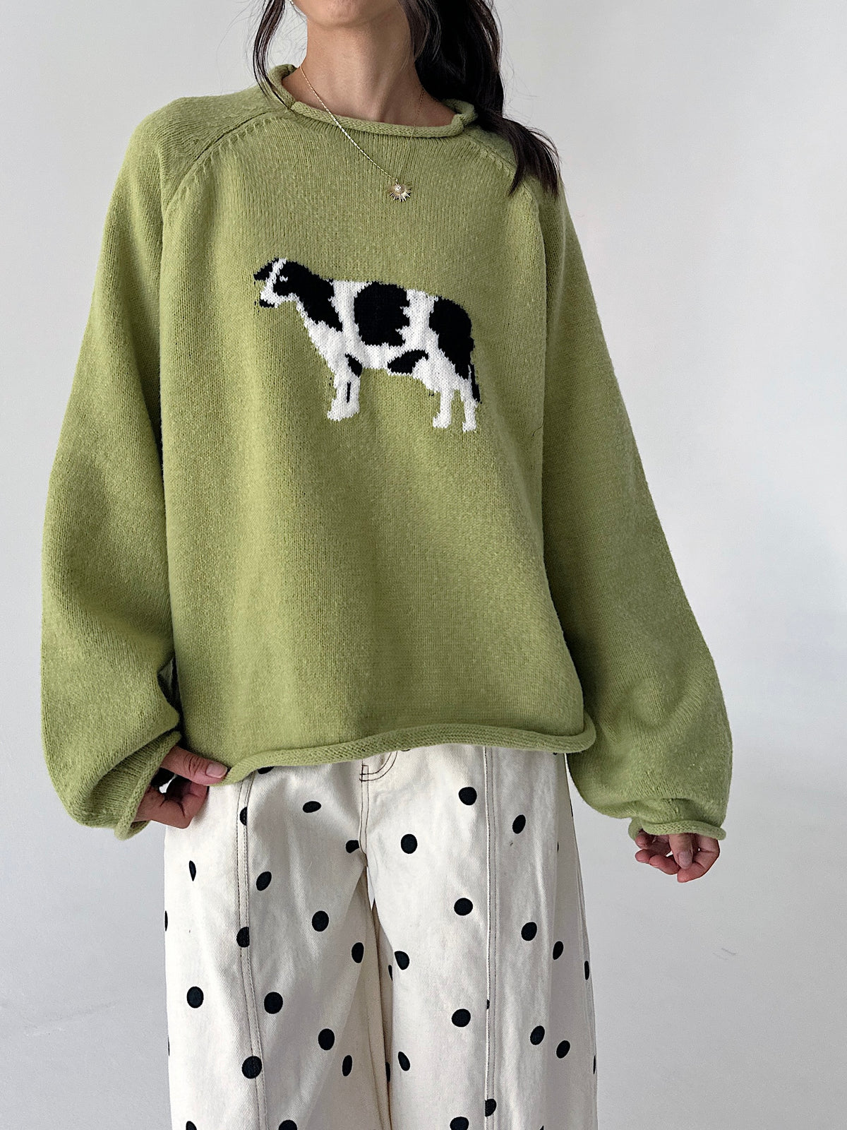 A person wearing an oversized sage green knit sweater with a black-and-white cow motif on the chest, paired with white pants with black polka dots, against a plain light gray background.