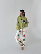 A woman stands barefoot against a plain light gray studio background, wearing a loose sage green sweater with a cow applique and white crocheted pants decorated with colorful granny-square patterns.