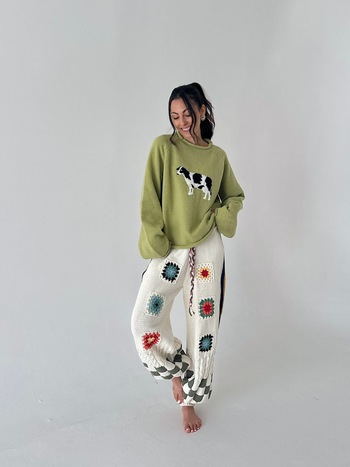 A woman stands barefoot against a plain light gray studio background, wearing a loose sage green sweater with a cow applique and white crocheted pants decorated with colorful granny-square patterns.