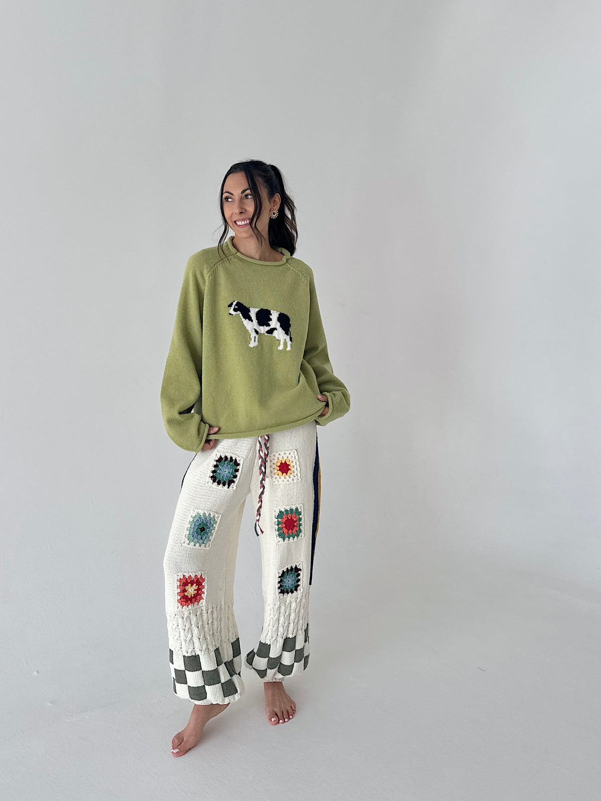 A woman stands barefoot in a studio wearing a light olive green crewneck sweater with a cow graphic and white crochet wide-leg pants with colorful floral motifs and black-and-white checkered cuffs.