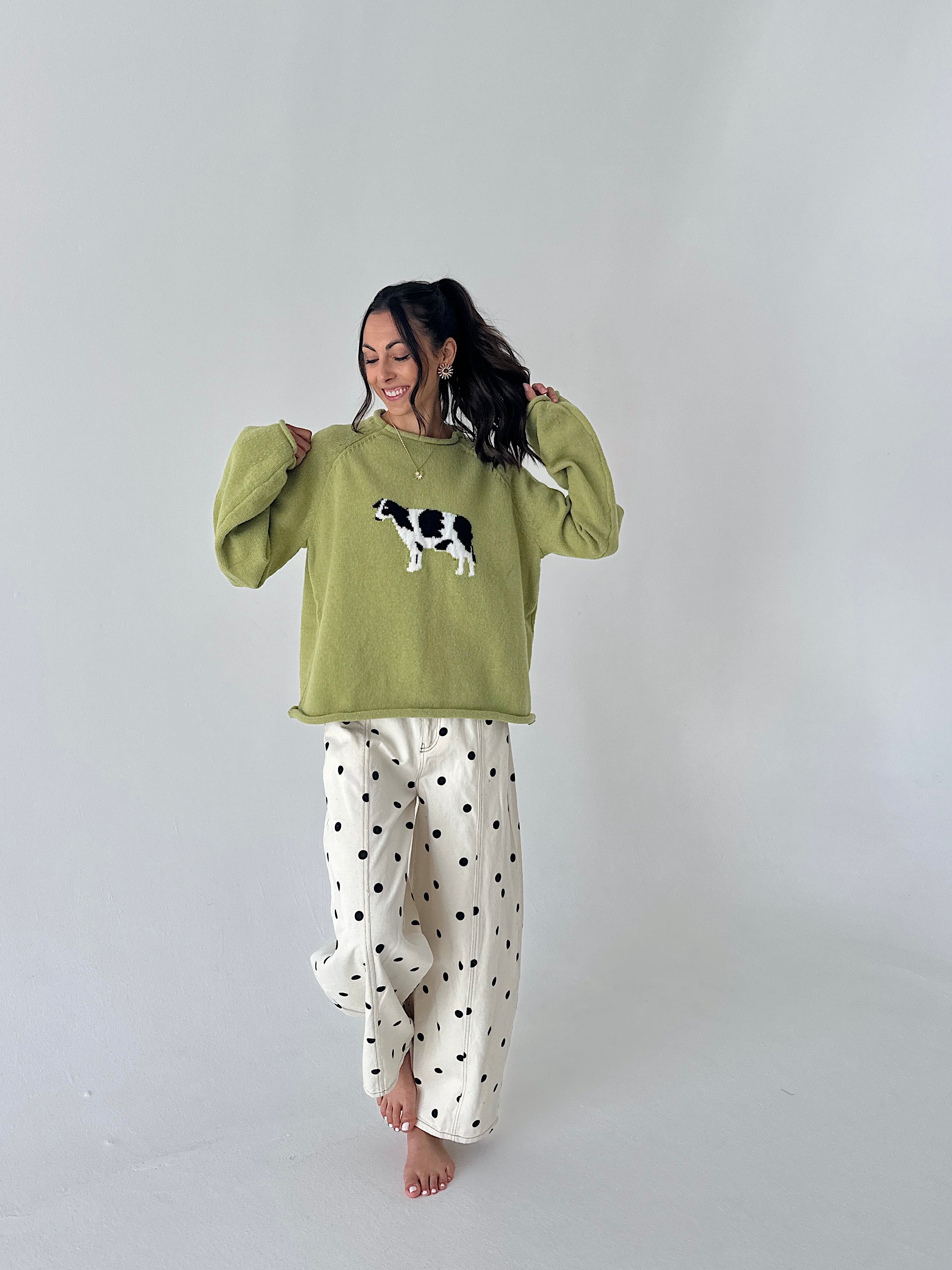 A woman modeling a green long-sleeve sweater with a black and white cow graphic on the front, paired with cream wide-leg pants featuring black polka dots, against a plain light gray studio background.
