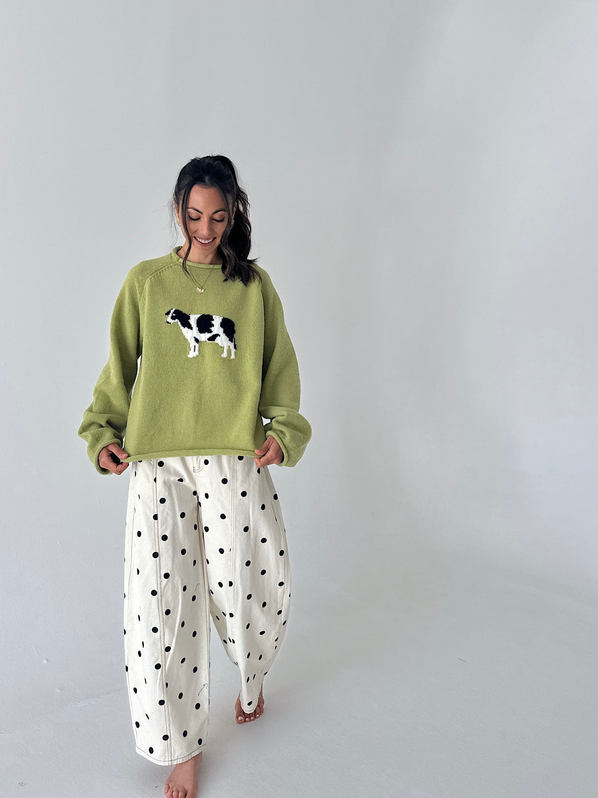 A woman modeling a light olive green crewneck sweater with a black and white cow graphic on the chest, paired with white wide-leg pants featuring black polka dots, set against a plain light gray studio background.