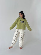 A woman modeling a cozy sage green crewneck sweater with a black and white cow applique on the chest, paired with white wide-leg pants featuring black polka dots, set against a plain light gray studio background.