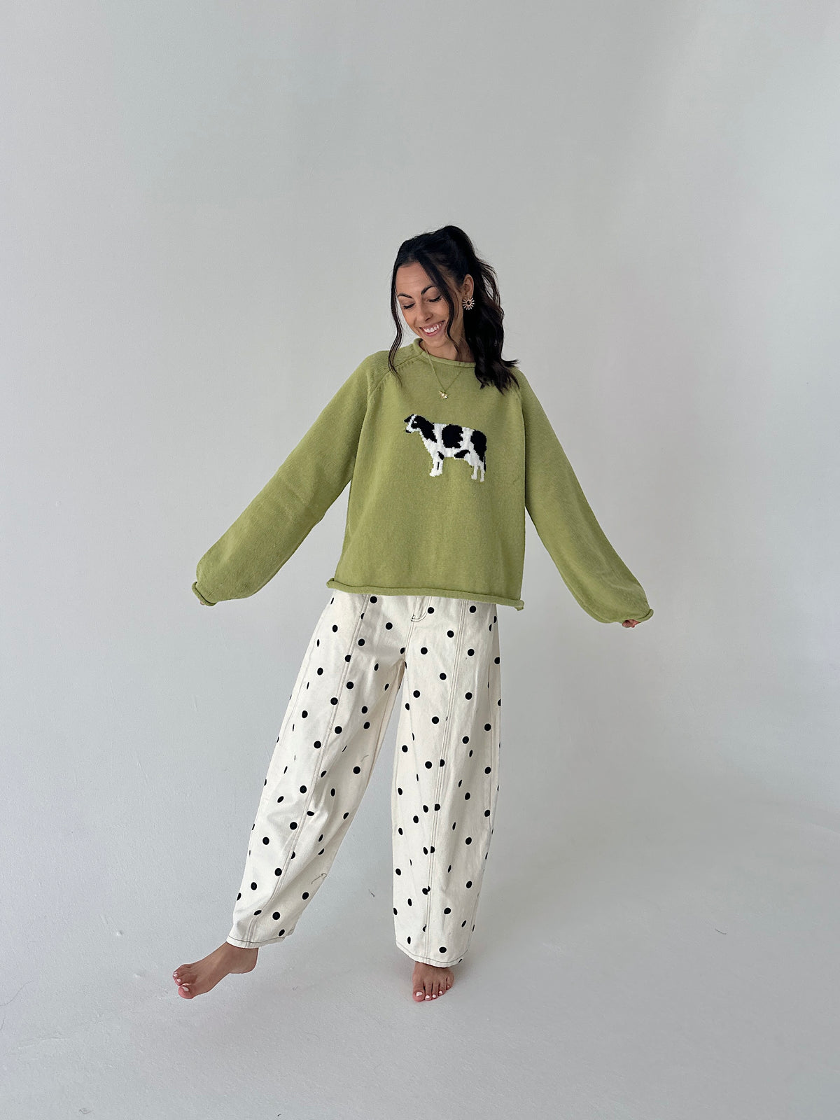 A woman modeling a cozy sage green crewneck sweater with a black and white cow applique on the chest, paired with white wide-leg pants featuring black polka dots, set against a plain light gray studio background.