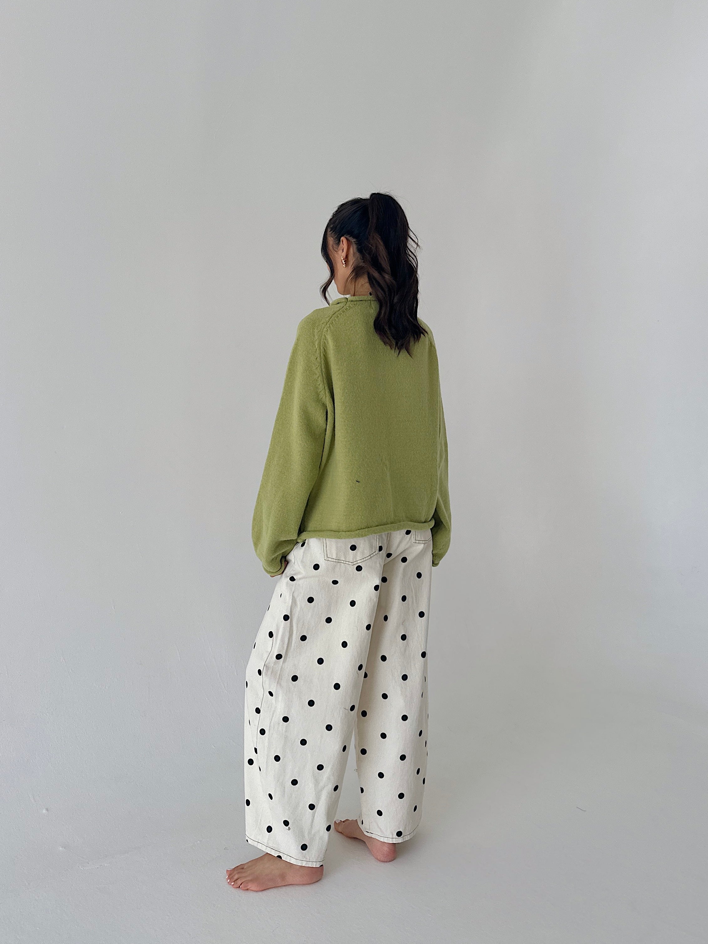 A woman standing with their back to the camera, wearing an olive green knit sweater and white wide-leg pants with black polka dots, set against a plain light gray studio background.