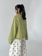 A woman shown from the back wearing a light olive green knit sweater and a white skirt with black polka dots, standing against a plain light gray wall.