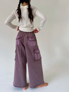 A person shown from the back wearing wide-leg mauve-purple cargo pants and a cream and gray striped long-sleeve shirt, standing barefoot against a plain light gray studio background.