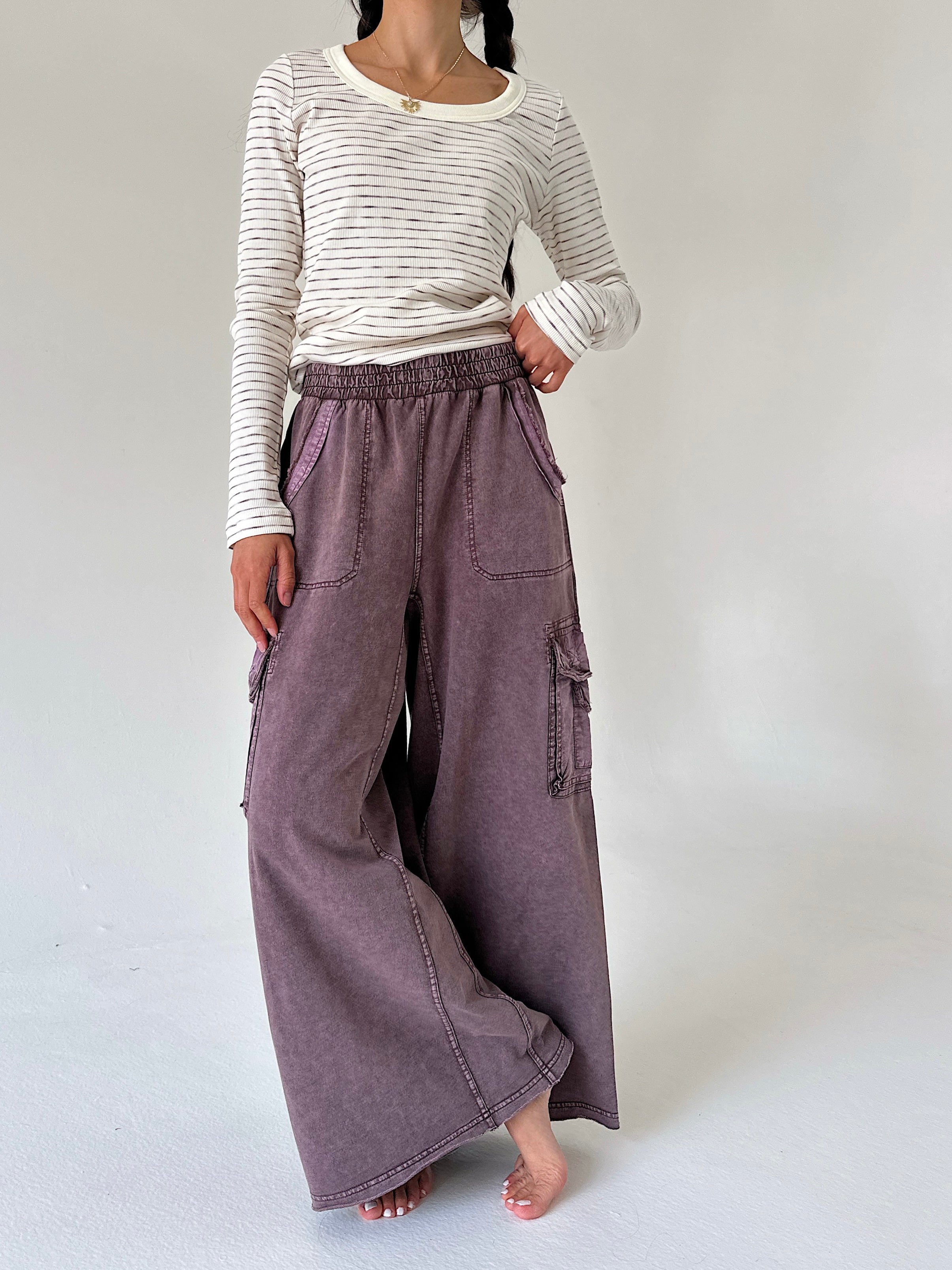 A model wearing a cream and gray striped long-sleeve shirt with wide-leg, wide-cuffed mauve purple cargo pants, standing barefoot against a light gray studio background.