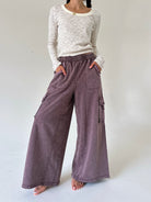 A model wearing wide-leg, cargo sweatpants in a washed mauve-purple color, paired with a cream and gray striped long-sleeve top, standing against a plain light gray studio background.