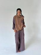 A woman stands barefoot against a plain light gray studio background, wearing a brown and mauve-purple patchwork hoodie with matching wide-leg cargo pants.