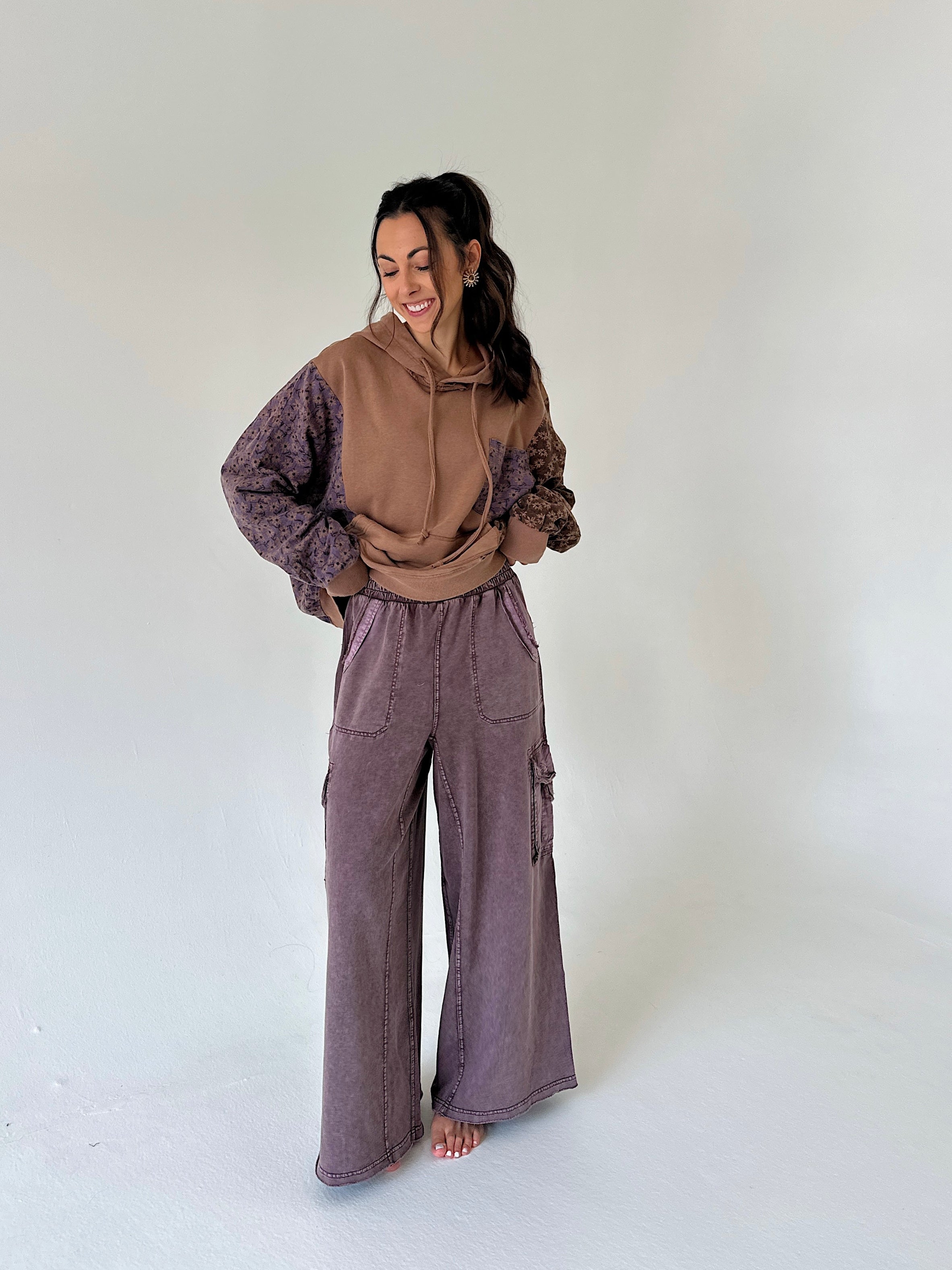 A woman modeling a two-tone brown and purple hoodie with patterned sleeves, paired with wide-leg purple cargo pants, standing against a plain light gray studio background.