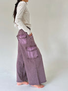 A person shown from the side wearing a cream and gray striped long-sleeve top and wide-leg mauve-purple cargo pants, standing barefoot against a plain light gray studio background.