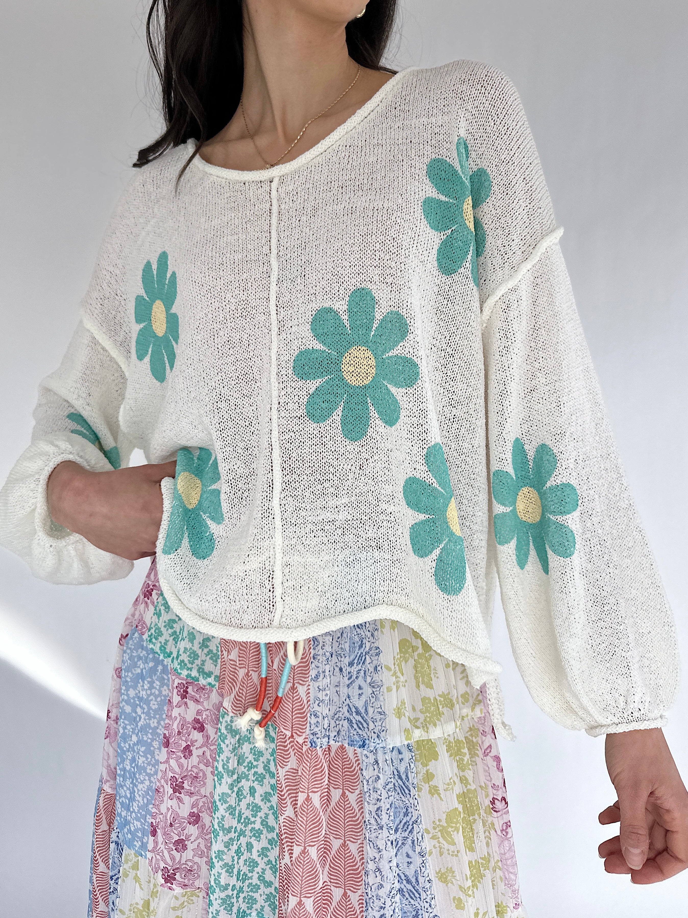 Woman wearing a white top with green floral patterns and colorful pants on a plain background