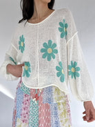 Woman wearing a white top with green floral patterns and colorful pants on a plain background