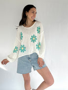 Woman wearing a white sweater with green floral patterns and denim shorts on a plain background