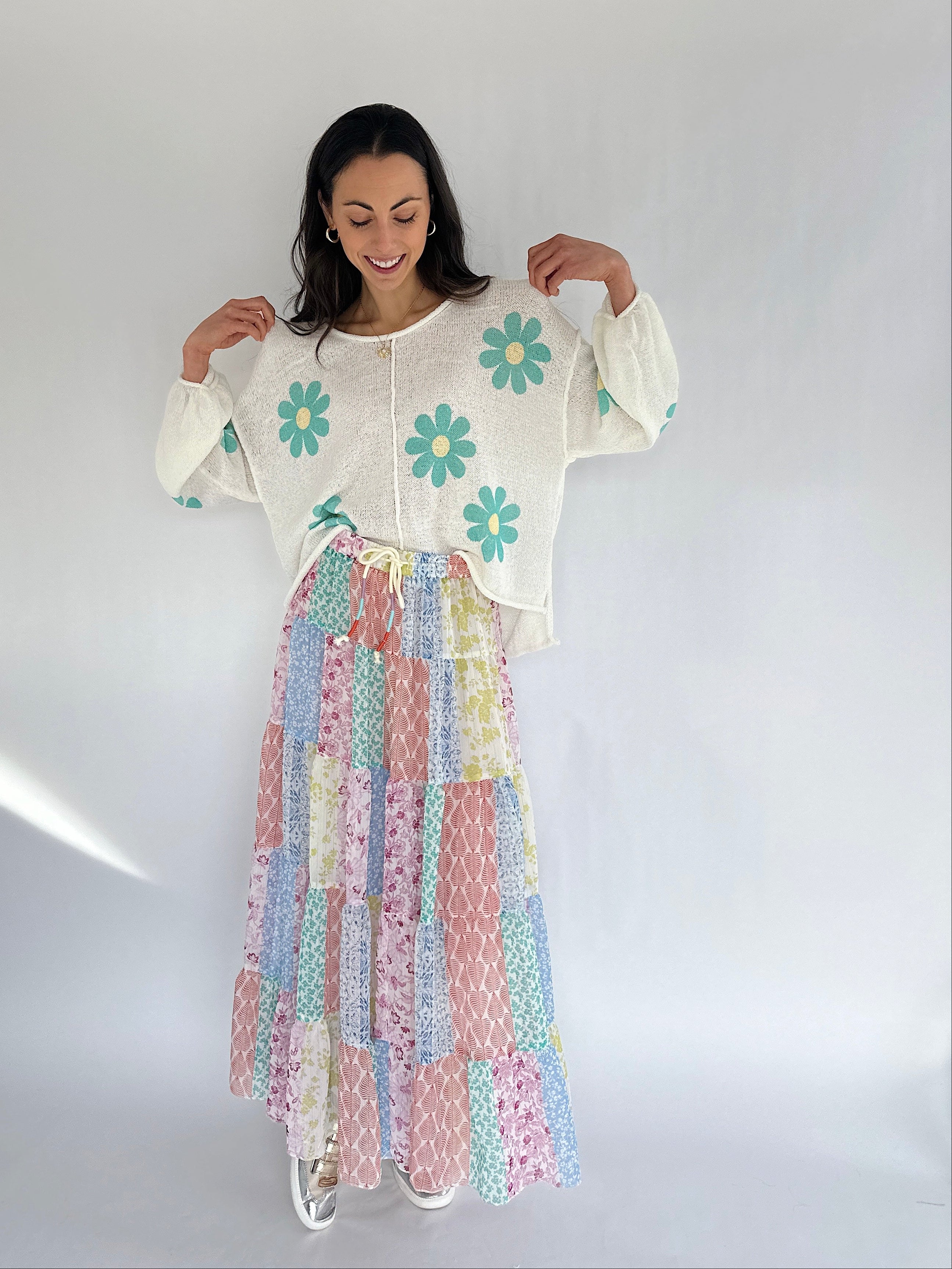 Woman wearing a white knit sweater with floral patterns and a colorful skirt against a white background