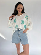 Woman wearing a white sweater with teal floral patterns and denim shorts on a plain background