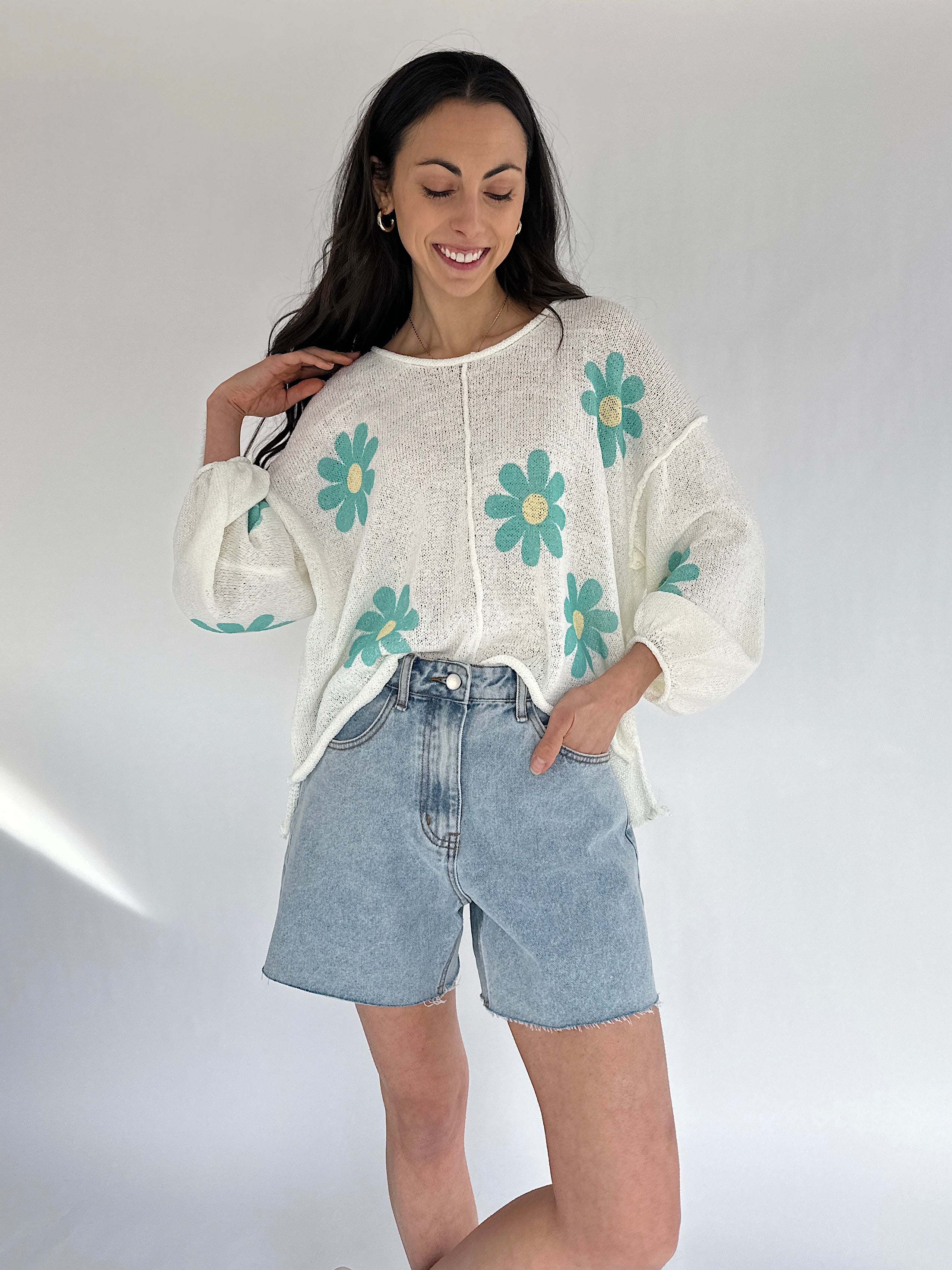 Woman wearing a white sweater with teal floral patterns and denim shorts on a plain background