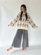 woman wearing a cream sweater with dancing skeletons and ash grey wide leg sweatpants in a studio setting