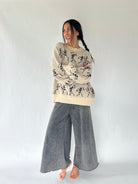 woman standing with her arms crossed wearing a cream sweater with dancing skeletons all over and a pair of wide leg sweatpants in an ash color