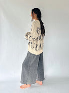 woman wearing a sweater with dancing skeletons and a pair of grey wide leg pants standing from the side in a studio setting