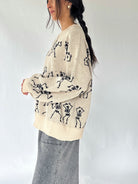 woman standing from the side wearing a cream sweater with dancing skeletons all over