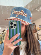 Person wearing a denim cap with 'Daughter' text, holding a striped phone case.