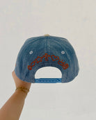 Denim cap with embroidered design held by a hand against a plain background