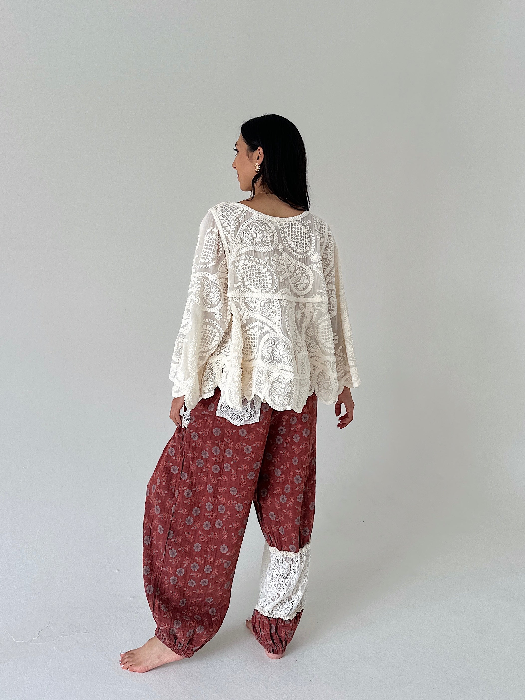 A woman stands barefoot against a plain light gray studio background, shown from the back. She is wearing a cream lace long-sleeve top over loose rust-red patterned harem-style pants with white lace cuffs.
