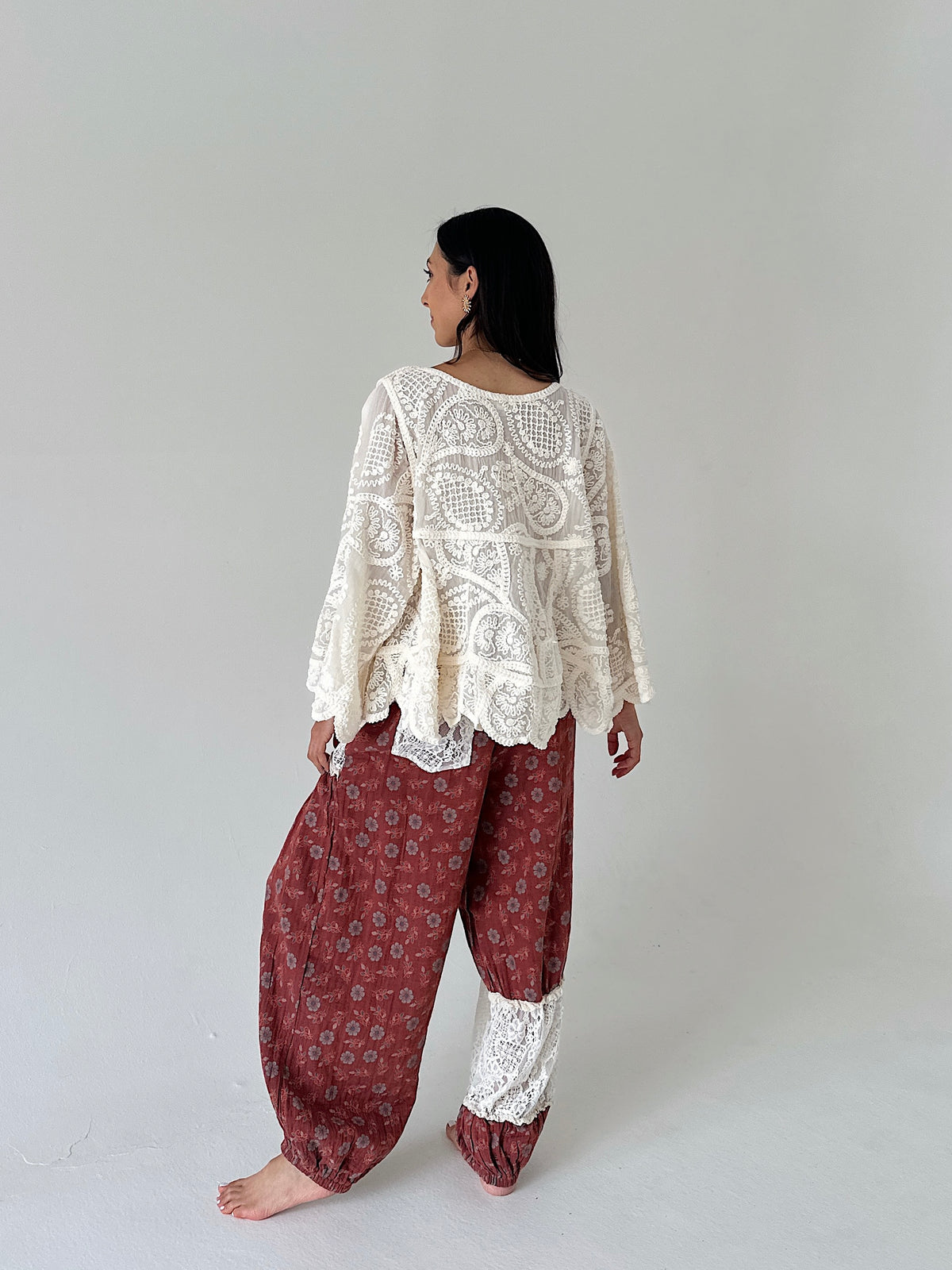A woman stands barefoot against a plain light gray studio background, shown from the back. She is wearing a cream lace long-sleeve top over loose rust-red patterned harem-style pants with white lace cuffs.