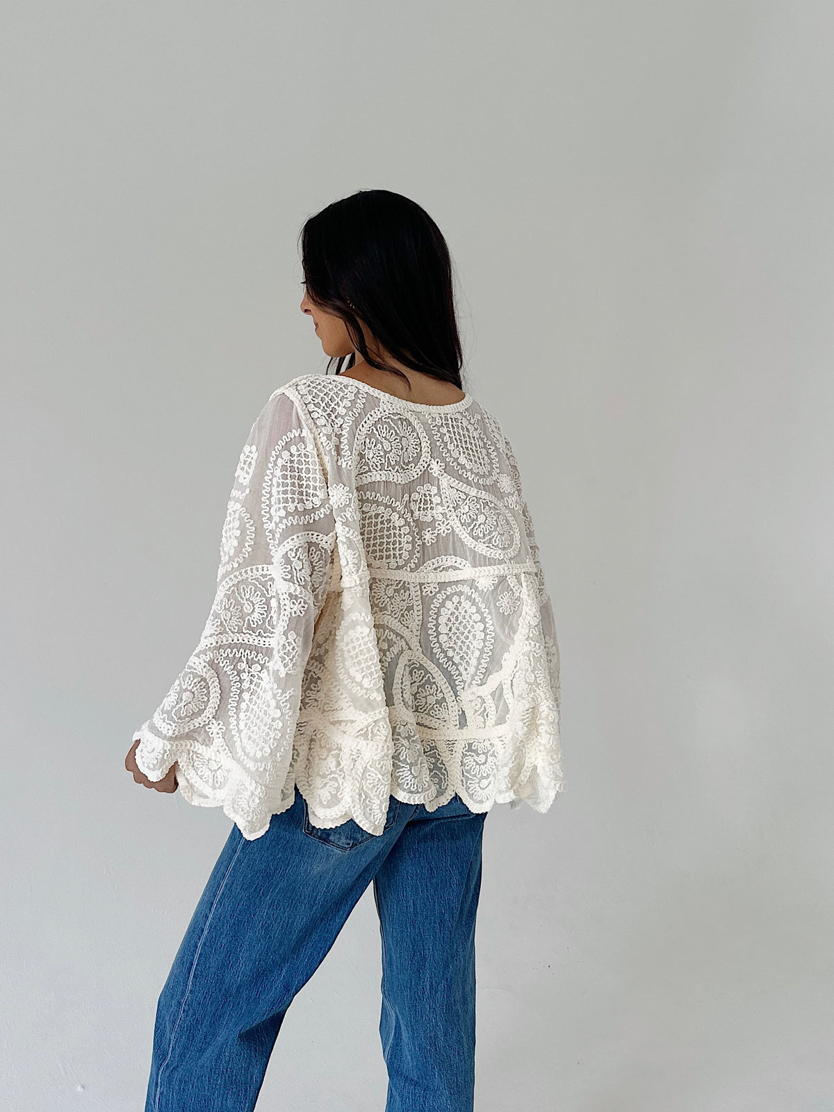 A woman shown from the back wearing a cream-colored, long-sleeved lace top and blue jeans against a plain light gray background.