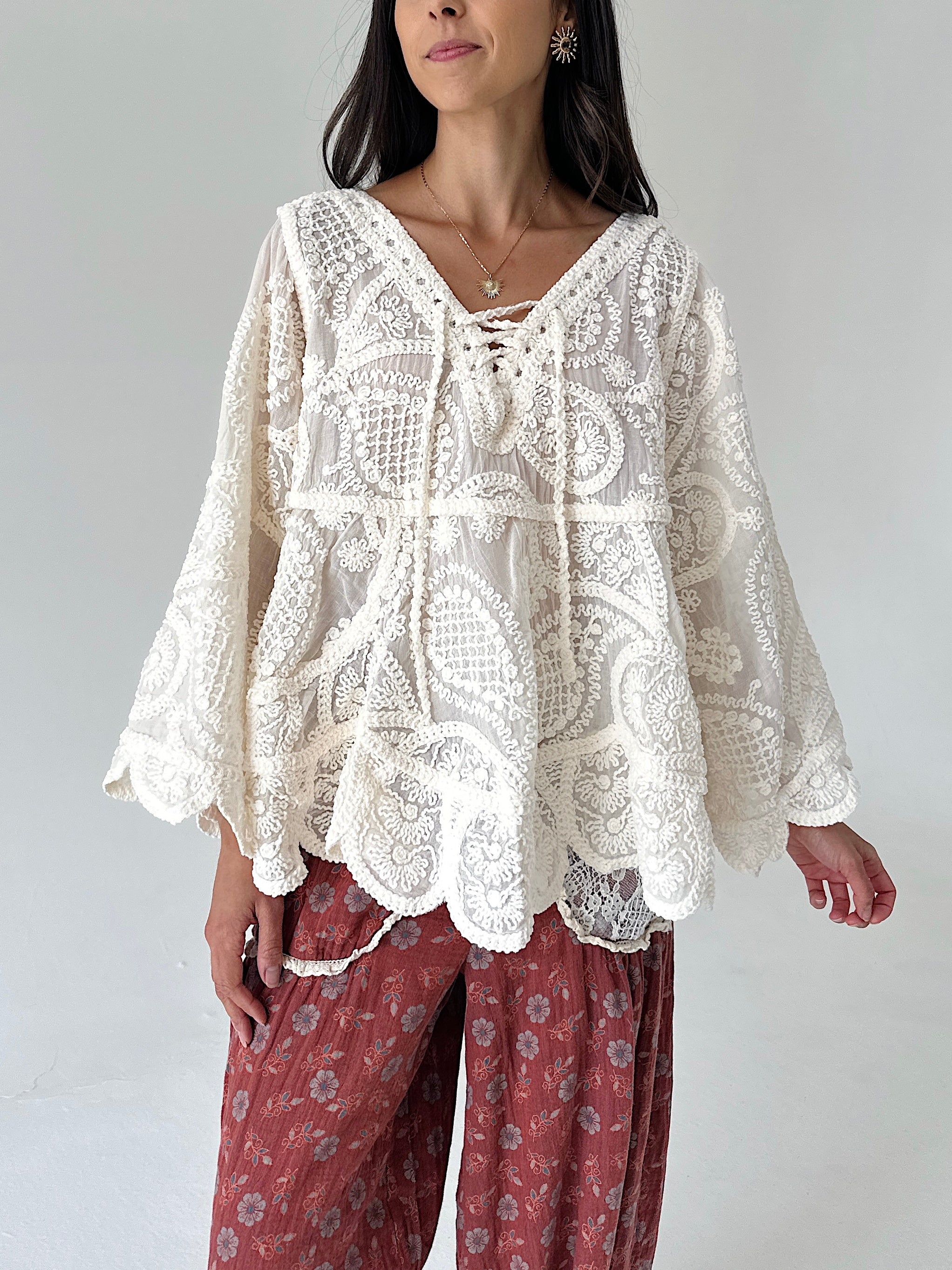 A woman wearing a cream-colored, long-sleeved lace top with a lace-up neckline and scalloped edges, paired with rust-colored floral pants, against a plain light gray background.