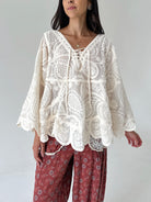 A woman wearing a cream-colored, long-sleeved lace top with a lace-up neckline and scalloped edges, paired with rust-colored floral pants, against a plain light gray background.