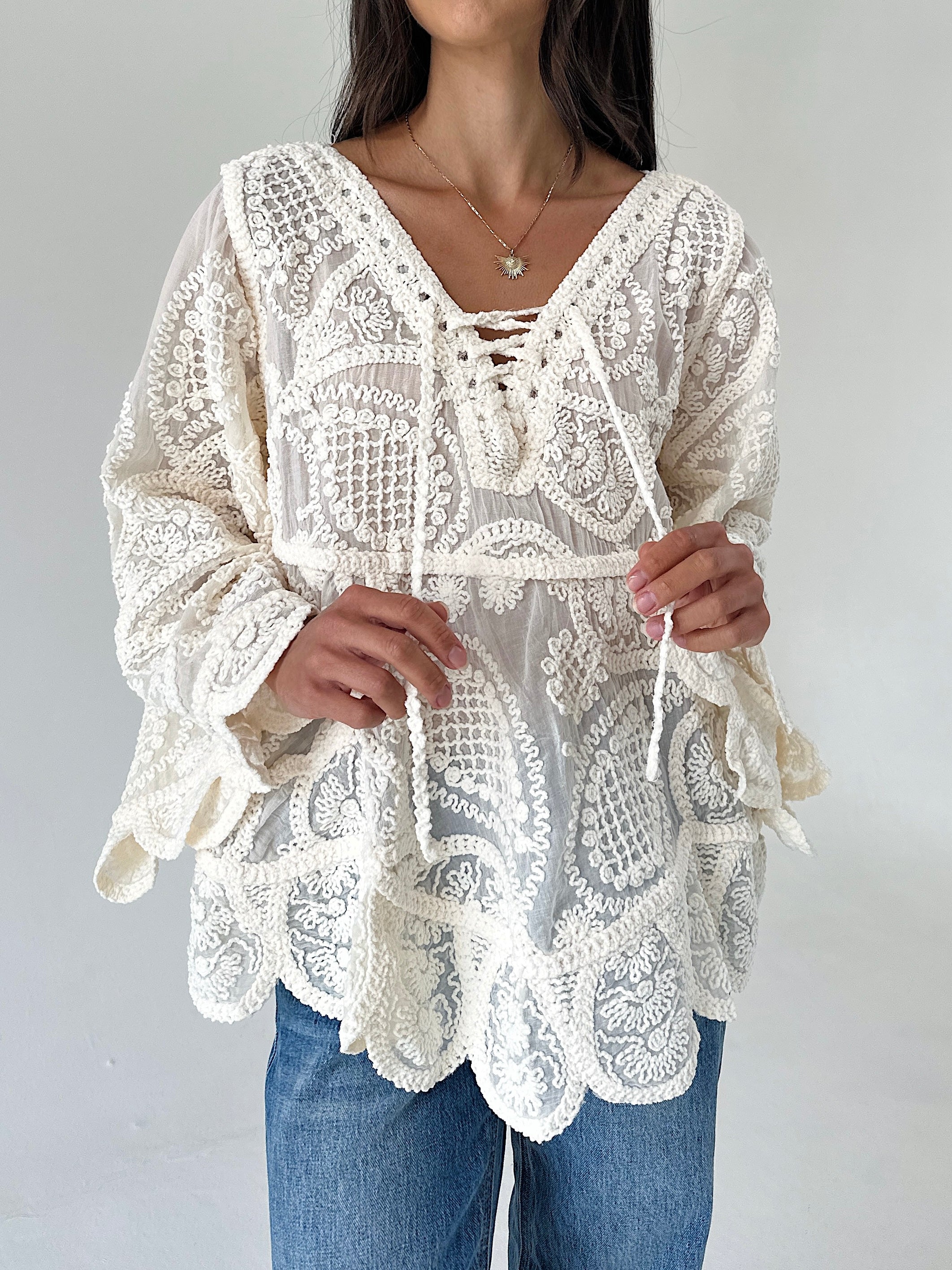 A woman wearing a cream-colored lace blouse with long bell sleeves, a V-neckline with tie-front details, and a scalloped hem, paired with blue jeans against a plain light gray background.
