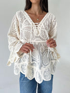 A woman wearing a cream-colored lace blouse with long bell sleeves, a V-neckline with tie-front details, and a scalloped hem, paired with blue jeans against a plain light gray background.