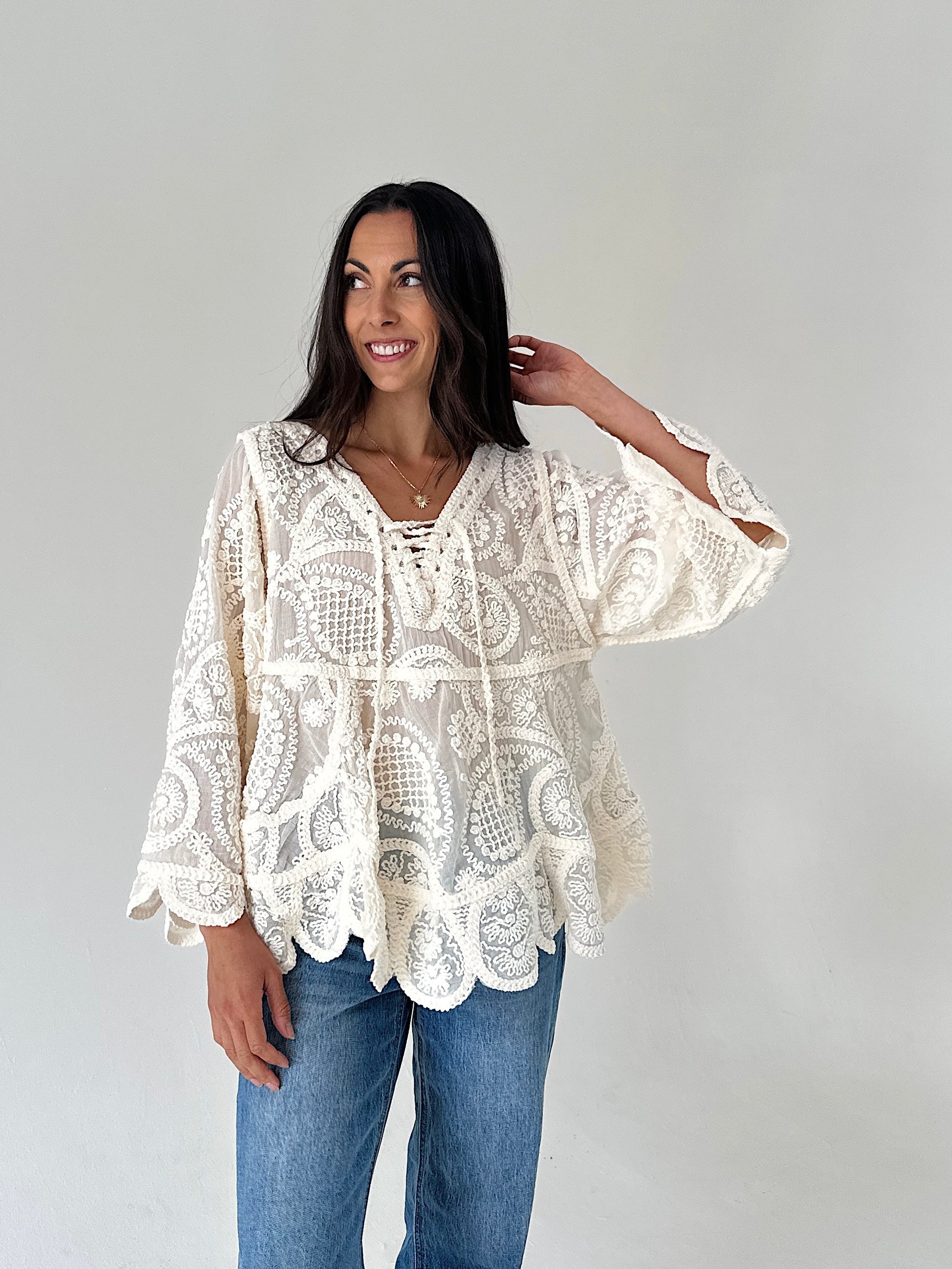 A woman modeling a cream lace bohemian-style top with blue jeans, standing against a plain light gray studio background.
