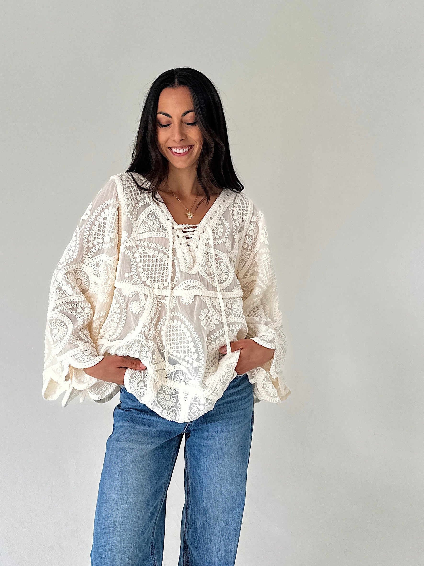A woman wearing a cream-colored crochet lace long-sleeve blouse with blue jeans, standing against a plain light gray background.